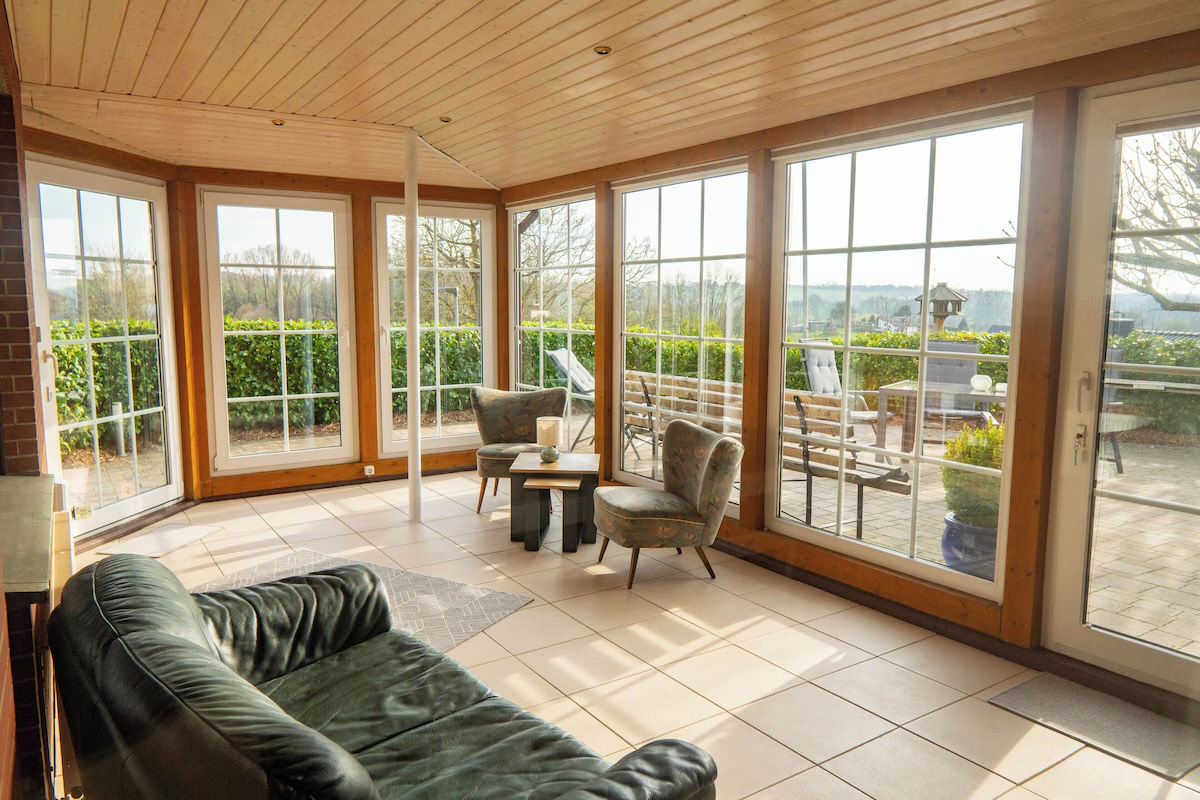 Sonniger Wintergarten mit Holzdecke, umlaufender Fensterfront, Sesseln und Blick auf Terrasse und Felder